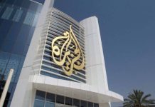 This image displays the headquarters building of Al Jazeera Media Network, located in Doha, Qatar. The facade features the network's iconic gold calligraphy logo set against modern white panels and reflective glass.