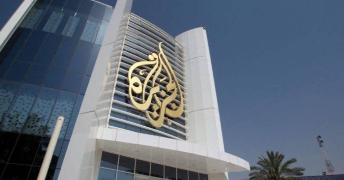 This image displays the headquarters building of Al Jazeera Media Network, located in Doha, Qatar. The facade features the network's iconic gold calligraphy logo set against modern white panels and reflective glass.