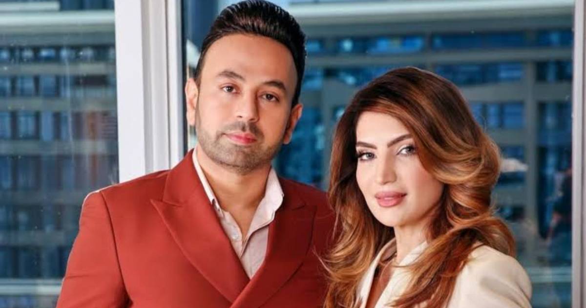 Satish Sanpal wearing a burnt orange double-breasted suit jacket standing next to his wife, Tabinda Sanpal, wearing a cream blazer in front of a high-rise window.