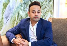 Entrepreneur and media personality Satish Sanpal sitting on a brown sofa wearing a navy blue blazer over an open-collar white shirt, with a marbled wall in the background.