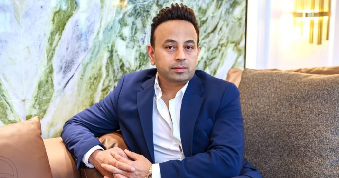 Entrepreneur and media personality Satish Sanpal sitting on a brown sofa wearing a navy blue blazer over an open-collar white shirt, with a marbled wall in the background.